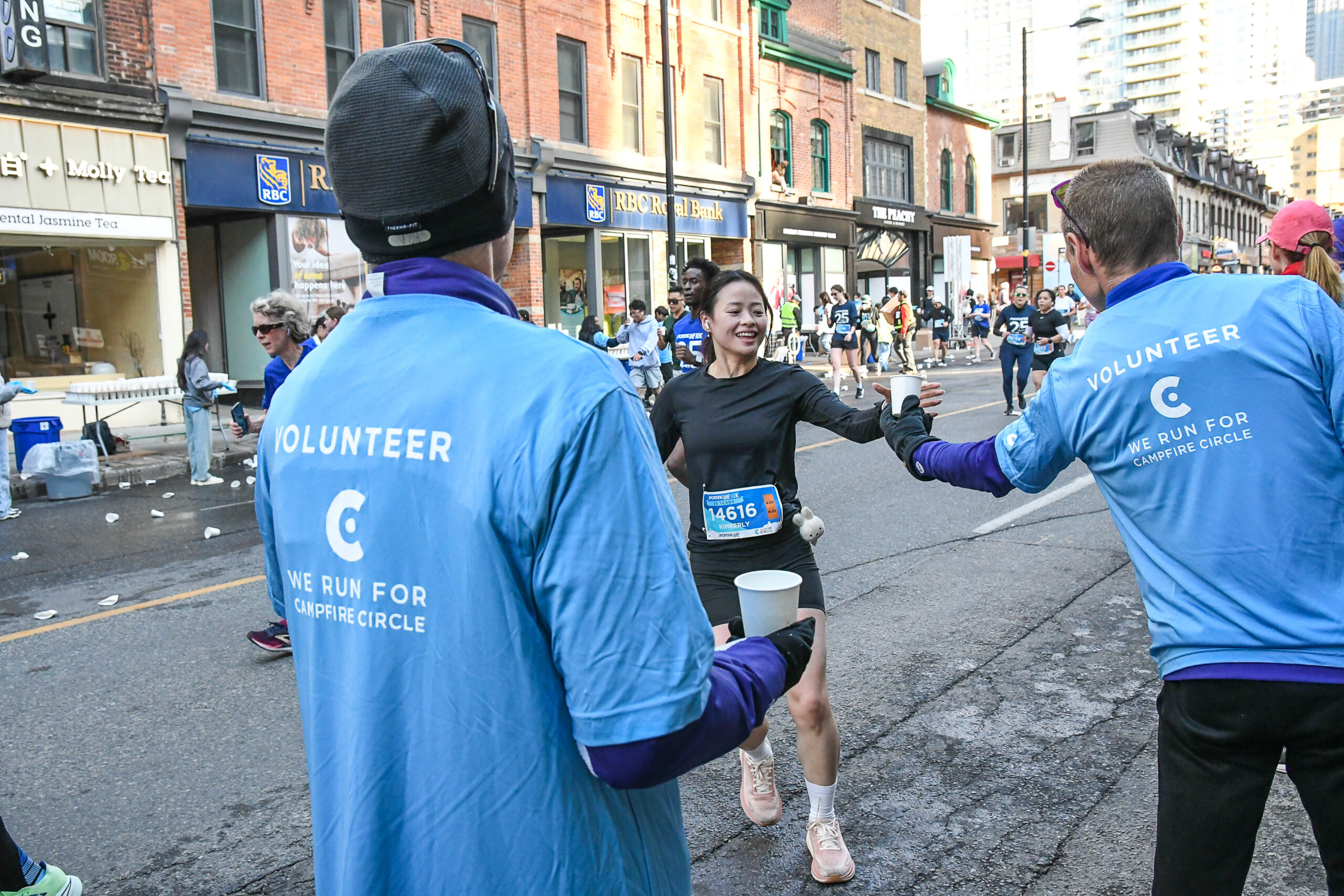 Smiling female runner reaches for water from volunteers wearing blue “We Run for Campfire Circle” shirts during the Sporting Life 10K in downtown Toronto, with cheering spectators and other runners in the background.
