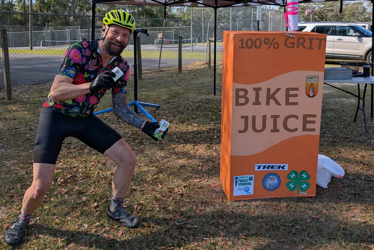 Cyclist posing with a giant “Bike Juice” carton prop at the Dirty Pecan Gravel Ride, under a Trek tent, celebrating grit and endurance.