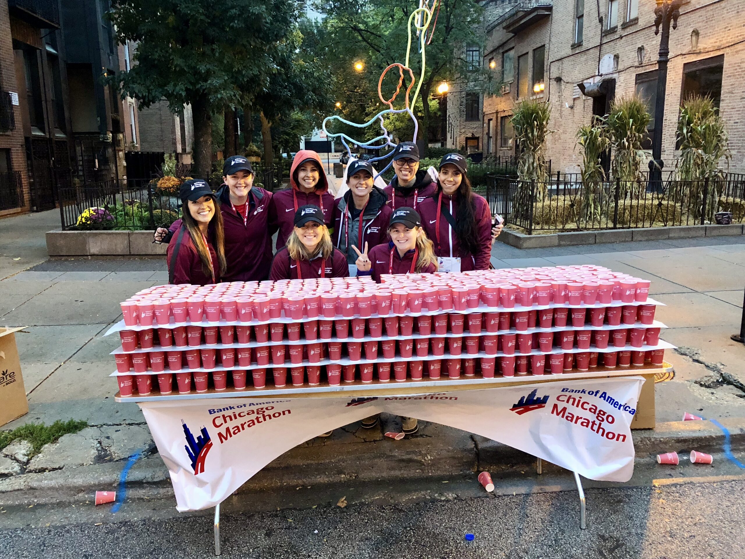 Group of volunteers in matching maroon jackets and hats smiling behind a table stacked with red water cups at the Chicago Marathon.