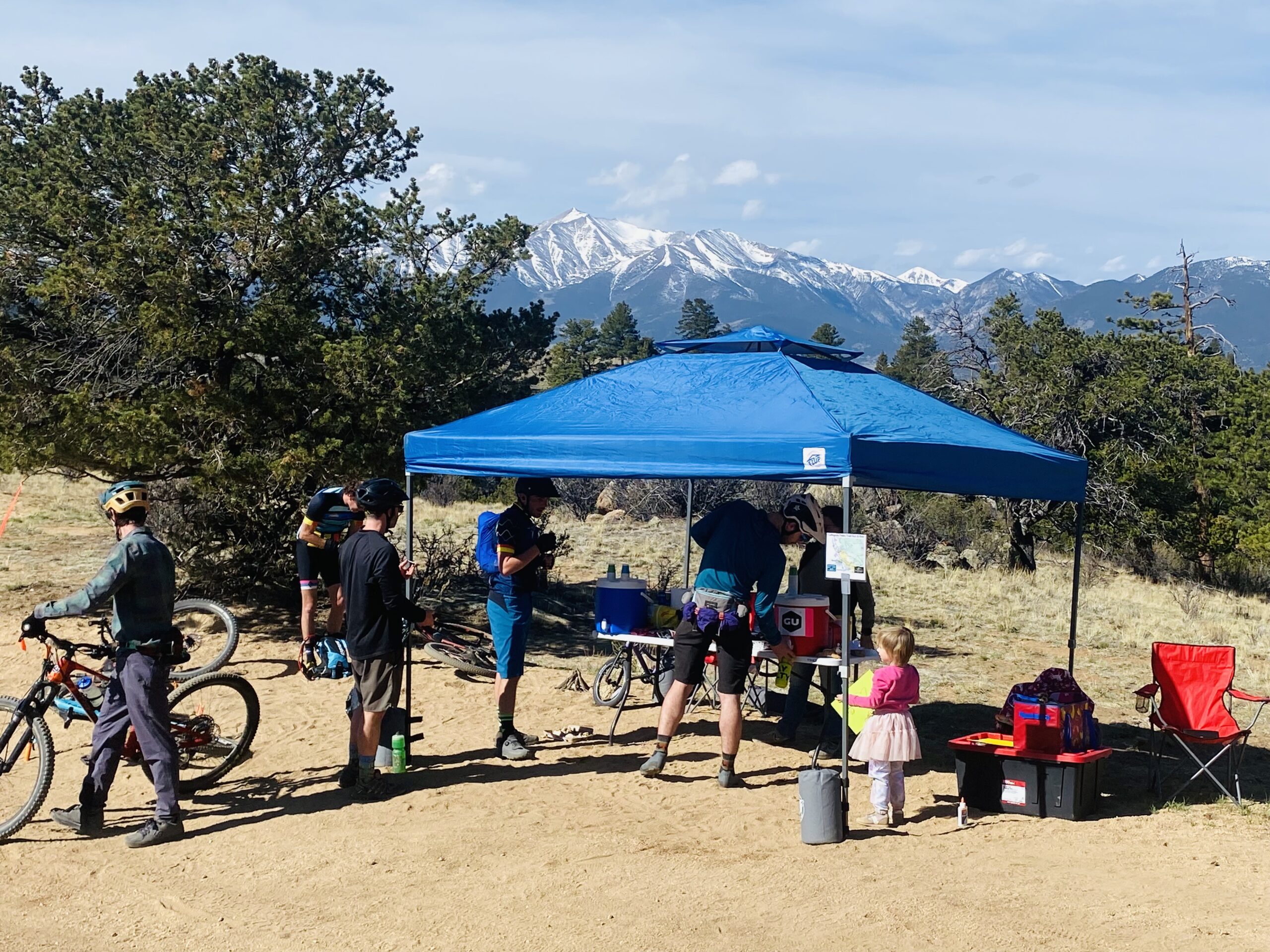Cyclists and volunteers gather at a packet pick-up tent during the Collegiate Peaks Trail Ride in Buena Vista, Colorado, with snowcapped mountains in the background.