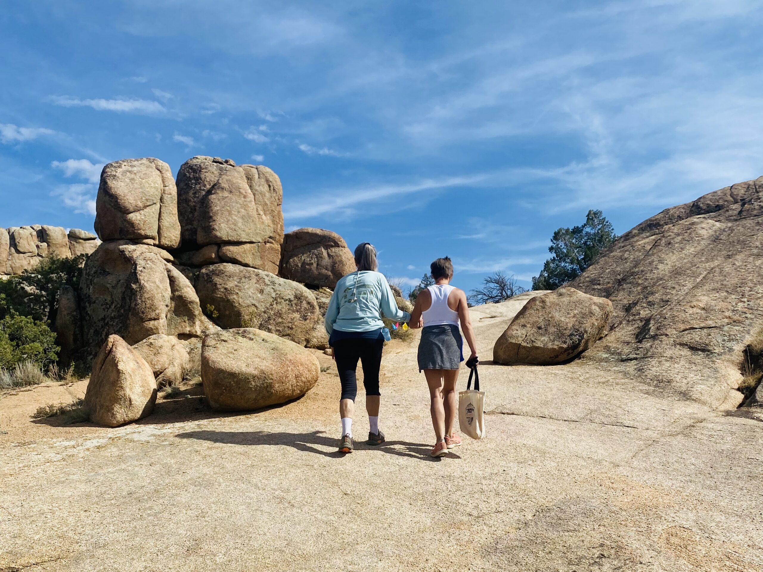 Two women walking together on a rocky trail under a bright blue sky, symbolizing community and support at endurance events.