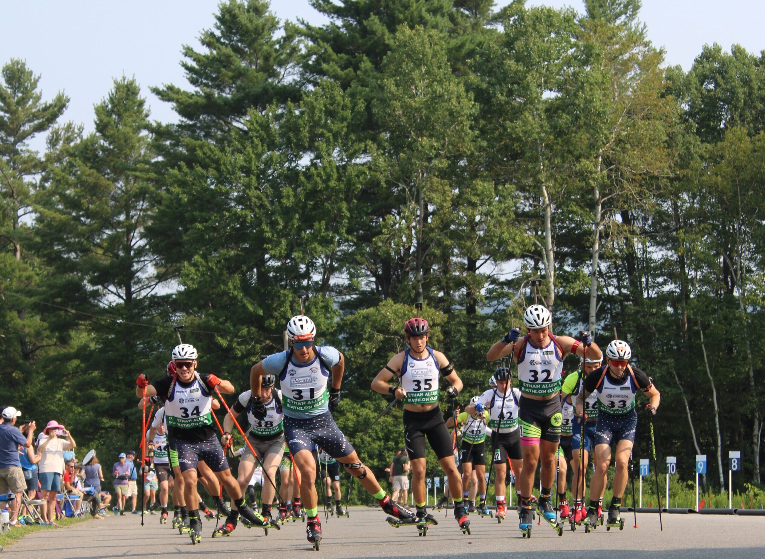 Biathletes racing on roller skis during the USBA Summer National Biathlon Championships, competing on a sunny day with a crowd watching.