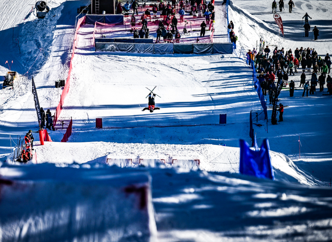 A freestyle moguls competitor goes inverted mid-air above the course at U.S. Freestyle Moguls Selections, with spectators lining the snow-covered venue on a clear winter day.