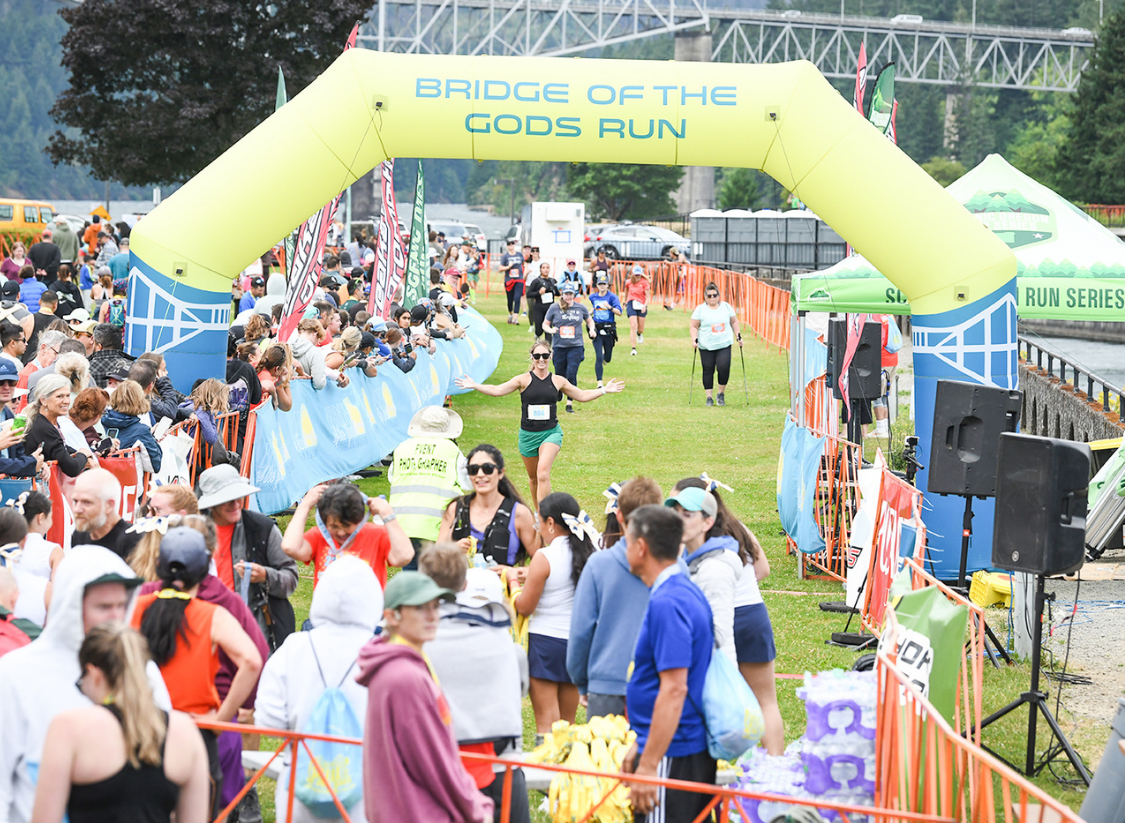 Finish line scene at the Bridge of the Gods Run, with runners crossing under the finish arch as spectators cheer from both sides of the course.
