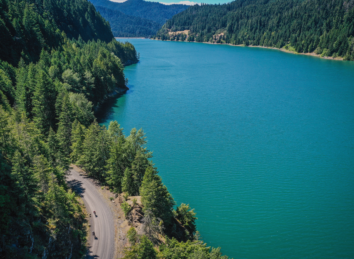 Aerial view of cyclists riding along a gravel road beside a blue lake surrounded by forest in Bend, Oregon during Oregon Trail Gravel.