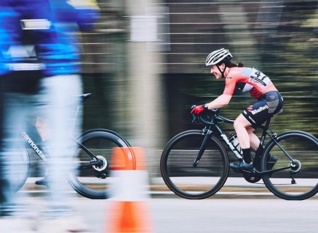 A smiling cyclist in a red BikeReg jersey races past orange cones during a criterium event, captured with a motion-blur effect.
