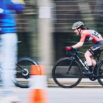 A smiling cyclist in a red BikeReg jersey races past orange cones during a criterium event, captured with a motion-blur effect.